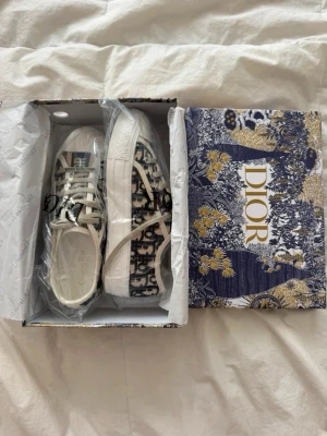 Dior Oblique sneakers i blått och vitt - Snygga Dior Oblique sneakers med klassiskt blått och vitt mönster samt vita snören. Skorna har lågt skaft och tydlig logga på plösen och sidan. Ovandelen är i canvas och sulan är vit med diskret mönster. Perfekta för dig som vill sticka ut med lyxig streetstyle.