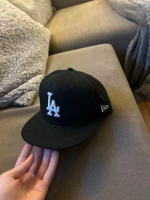 Svart LA New Era 59FIFTY keps - Snygg svart keps från New Era med LA-logga broderad i vitt framtill. Klassisk 59FIFTY-modell med rak skärm och broderade ventilationshål. Märkeslogga på sidan och insida med 59FIFTY-band. Perfekt för dig som gillar streetwear och sportig stil.