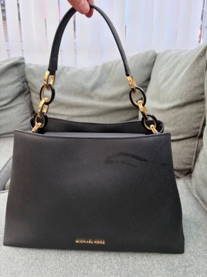Svart handväska från Michael Kors - Stilren svart handväska från Michael Kors i slätt läder med guldfärgade metalldetaljer och kedjehandtag. Väskan har en klassisk, fyrkantig form och logotypen i guld framtill. Perfekt för dig som gillar en elegant och tidlös look.