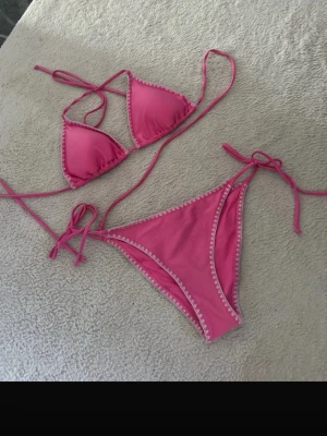 Rosa bikini från Gina Tricot - Säljer en snygg rosa bikini från Gina Tricot med vita dekorativa sömmar längs kanterna. Både överdel och underdel har knytband i sidorna och triangelformad topp. Perfekt för lata dagar på stranden eller poolen. Helt ny aldrig använd