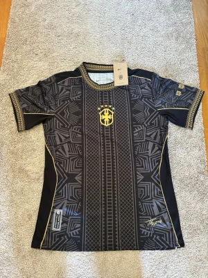 Svart och guld fotbollströja Nike - Snygg svart fotbollströja från Nike med guldiga detaljer och geometriskt mönster. Tröjan har korta ärmar, rund hals och guldiga sömmar längs sidorna. Märkt med 'Authentic Dols Apparel Product' och har ett sportigt tryck framtill.