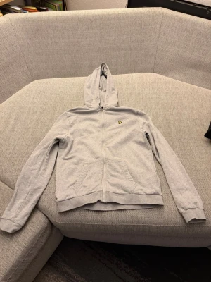 Grå hoodie från Lyle & Scott - Snygg grå hoodie från Lyle & Scott med dragkedja framtill och klassisk huva. Tröjan har två fickor och den ikoniska gula loggan på bröstet. Tillverkad i mjukt bomullsmaterial som är skönt att ha på sig. Perfekt för en avslappnad stil.