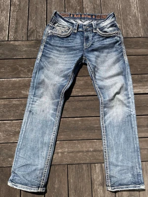 *SUPER RARE* sällsynta eftertraktadeRock Revival ljusblå jeans - Säljer ett par ljusblå jeans från Rock Revival med slitna detaljer och kontrastsömmar. Jeansen har broderade bakfickor med grått mönster och tydliga tvättade effekter. Klassisk femficksmodell och raka ben. Perfekta för dig som gillar streetwear och detaljer.