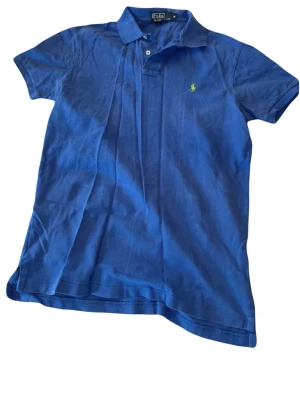 Blå pikétopp från Ralph Lauren - Blå pikétopp från Ralph Lauren med klassisk krage, korta ärmar och två knappar framtill. Toppens ikoniska logga är broderad i gult på bröstet. Tillverkad i mjuk bomull för en bekväm känsla och stilren look.