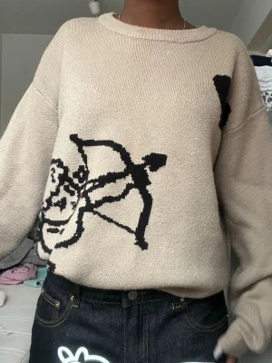 Beige stickad tröja med svart tryck - Beige stickad tröja med svart grafiskt tryck av en bågskytt på framsidan och texten 'I WOULD RATHER DIE THAN NOT LOVE' på ryggen. Tröjan har avslappnad passform, rund halsringning och långa ärmar. Perfekt för dig som gillar streetwear vibes.