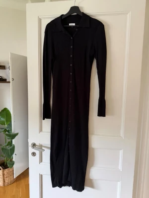 Svart ribbad långklänning med knappar - Lång svart ribbad klänning med knappar hela vägen framtill och krage. Klänningen har lång ärm och en smickrande, figurnära passform. Perfekt för dig som gillar stilrena och minimalistiska plagg.