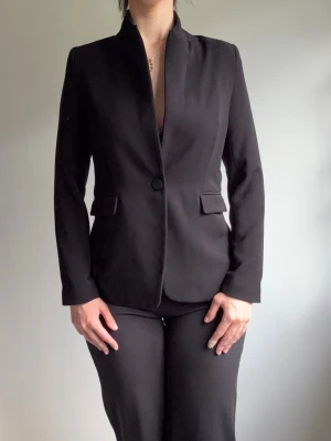 Svart figursydd kavaj  - Stilren svart kavaj med figurnära passform och modern ståkrage. Kavajen har en knapp framtill, två fickor med lock och långa ärmar. Perfekt för en clean och snygg look. Materialet ser ut att vara ett slätt tyg, troligen polyesterblandning.