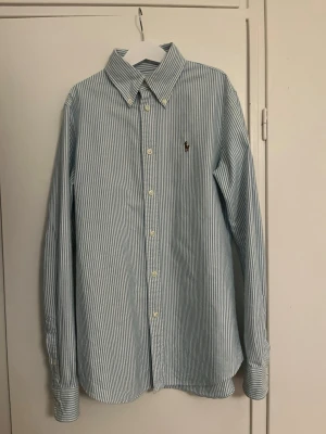 Randig skjorta från Ralph Lauren - Klassisk skjorta från Ralph Lauren med smala blå och vita ränder. Skjortan har lång ärm, knappar framtill och den ikoniska broderade loggan på bröstet. Tillverkad i mjuk bomull.