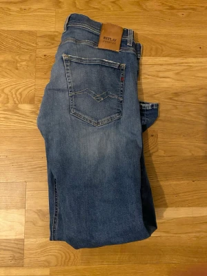 Blå Replay slim fit jeans - Säljer ett par blå slim fit jeans från Replay med klassisk femficksdesign och snygga slitningar på låret. Jeansen har en skinnpatch med Replay-logga bak i midjan och är tillverkade i mjukt denimtyg. Perfekta för en avslappnad och trendig look.