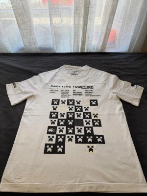 Vit Lacoste x Minecraft T-shirt - Säljer en vit T-shirt från Lacoste i samarbete med Minecraft. T-shirten har svart tryck med Minecraft-motiv och texten 'Crafting Together' på bröstet. Modellen är loose fit och har korta ärmar. Perfekt för dig som gillar gaming och streetwear.