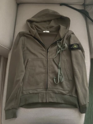 Grön hoodie från Stone Island - Säljer en snygg gr hoodie från Stone Island. Den har en dragkedja framtill och en klassisk huva. På ärmen finns det ikoniska Stone Island-märket. Den är i bra skick. Passar både till vardags och lite mer avslappnade still. Man får med snören. Skicka vid frågor. 