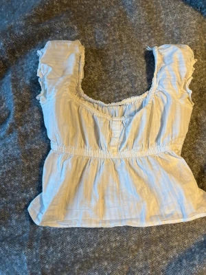 Vit volang topp med knappar - Söt vit topp med volangdetaljer och korta ärmar. Toppen har en smockad midja, knappar framtill och en luftig, lätt struktur. Perfekt för varma dagar och ger en romantisk vibe till din outfit.