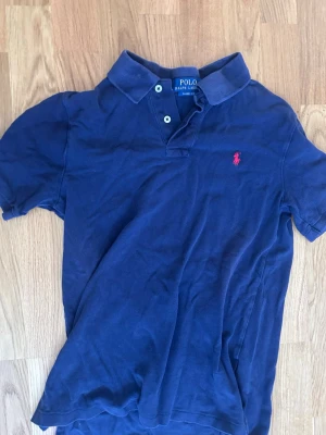 Marinblå pikétröja från Polo Ralph Lauren barnstorlek - Obs! Barnstorlek Klassisk marinblå pikétröja från Polo Ralph Lauren med korta ärmar och krage. Tröjan har två knappar vid halsen och den ikoniska röda logotypen broderad på bröstet. Tillverkad i bomull för en bekväm känsla.