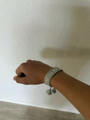 Dior armband - Grått armband från Christian Dior i textil med broderad logga och snygga tofsar. Armbandet har justerbar knytning så det passar de flesta handleder. Perfekt accessoar för att ge din look en exklusiv touch.