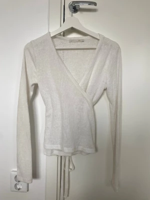 Vit omlottopp med knytband - Snygg vit omlottopp med långa ärmar och ribbad struktur. Toppen har knytband i midjan och v-ringad halsringning. Perfekt att styla med jeans eller kjol för en clean look.