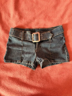 Mörkblå jeansshorts med bälte - Säljer ett par mörkblå jeansshorts med bredt matchande bälte och stor silverfärgad spänne. Shortsen har dekorativa sömmar framtill och en lågmidjad passform. Aldrig använda 