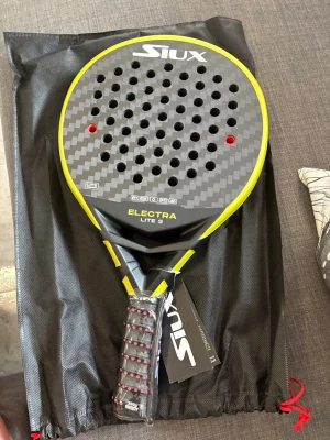 Siux Electra Lite 3 padelracket - Padelracket från Siux, modell Electra Lite 3. Racketen har en rund form med svart och gul design samt kolfiberyta. Greppet är svart med röda detaljer och racketen har flera hål för bättre kontroll. Perfekt för dig som vill ta ditt padelspel till nästa nivå.