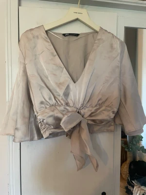 Champagnefärgad satin omlottopp från Zara - Snygg champagnefärgad omlottopp från Zara i glansigt satinmaterial. Toppen har v-ringning, trekvartsärm och en stor knytrosett i midjan som ger en elegant look. Perfekt för dig som vill sticka ut lite extra.