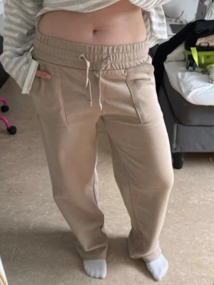 Beige mjukisbyxor med raka ben (Stella Low Waist)  - Säljer ett par beige mjukisbyxor med raka ben och bred resår i midjan. Byxorna har stora fickor framtill och en bakficka, samt dragsko i midjan för skön passform. Perfekta för chill dagar eller när du vill vara extra bekväm.