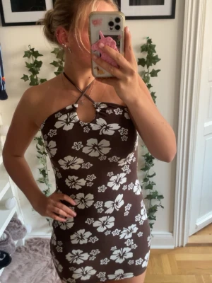 Brun blommig halterneckklänning - Supersnygg brun kortklänning med vita blommor och halterneck. Klänningen har en liten cut-out framtill och är figurnära med stretchigt material. Perfekt för sommardagar och när du vill sticka ut lite extra.