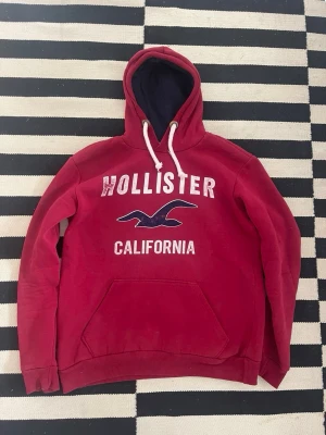 Röd Hollister hoodie med tryck - Snygg röd hoodie från Hollister med stort vitt och blått tryck på bröstet där det står 'Hollister California' och en våg. Mörkblå insida på huvan och vita dragsnören. Klassisk känguruficka framtill och ribbade muddar. Storlek L men är mer storlek M än L.