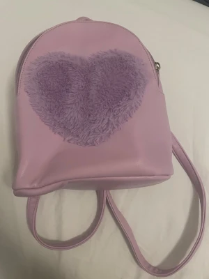 Ljuslila ryggsäck med fluffigt hjärta - Supergullig ljuslila ryggsäck med ett stort fluffigt lila hjärta på framsidan. Väskan har justerbara axelremmar och är gjord i ett slätt konstlädermaterial. Perfekt för dig som gillar söta detaljer och pastellfärger.