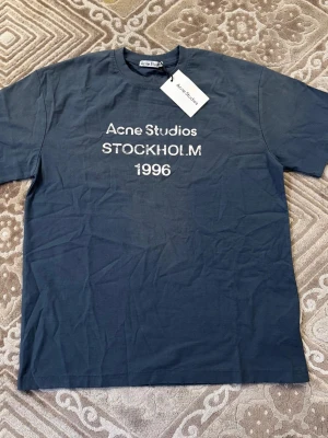 Blå Acne Studios t-shirt Stockholm - Helt ny och oanvänd T-shirt från Acne Studios med klassiska “Stockholm 1996”-trycket framtill. Snygg vintageinspirerad blågrå färg som ger en clean och avslappnad look. • Storlek: M (men passar mer som L) • Skick: helt ny / aldrig använd • Färg: blågrå, tvättad vintage-stil • Passform: lite oversized känsla Perfekt till vardags eller för en enkel streetwear-outfit. Skickas snabbt 🚚