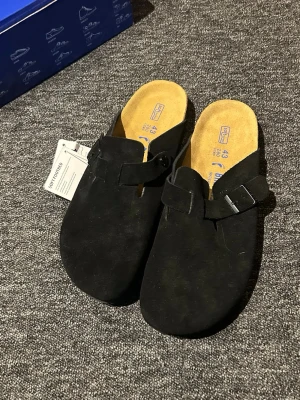 Svarta Birkenstock tofflor mocka - Säljer ett par svarta Birkenstock tofflor i mocka med justerbart spänne och mjuk innersula. Klassisk slip-in modell med ergonomisk fotbädd och snygg minimalistisk design. Perfekta för chill dagar hemma eller när du vill ha det bekvämt.
