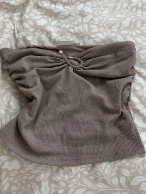 Beige offshoulder topp Kappahl - Trendig beige offshoulder topp från Kappahl i ribbad struktur. Toppen har en snygg detalj med pärla och ring framtill som ger en unik look. Perfekt att styla med höga jeans eller kjol för en cool outfit.