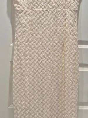 Beige spetsklänning axelbandslös - Axelbandslös beige klänning med vackert spetsmönster och figurnära passform. Klänningen har en elegant struktur och är perfekt för dig som gillar en stilren look. Materialet känns mjukt och följsamt mot kroppen.