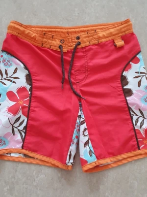 Badshorts - Säljer ett par färgglada badshorts med röd front, orange midja och benkant samt blommigt mönster i rosa, rött, blått och brunt på sidorna. Snörning i midjan och cool surfstil. Perfekta för stranden eller poolen! Finn en fläck.