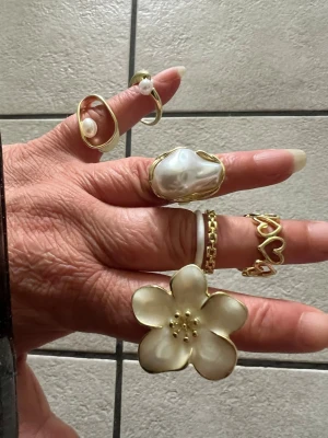 Guldiga ringar med pärldetaljer, 6 stycken  - Säljer ett set trendiga guldiga ringar i olika former, vissa med vita pärldetaljer och en stor blomma i pärlemor-look. Ringarna har unika designer med hjärtan, kedjemönster och öppna former. Perfekt för dig som vill mixa och matcha accessoarer.