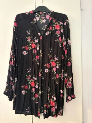 Svart blommig blus från bpc - Snygg svart blus från bpc med färgglada blommor i rosa, rött och vitt. Blusen har knappar framtill, v-ringning och långa ärmar. Materialet är mjukt och faller fint, perfekt för en trendig och avslappnad look. Storlek 44. 
