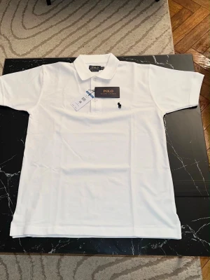 Vit pikétröja från Polo Ralph Lauren - Klassisk vit pikétröja från Polo Ralph Lauren med korta ärmar, krage och knappar framtill. Tröjan har den ikoniska broderade loggan i svart på bröstet och är tillverkad i bomull för en skön och luftig känsla.