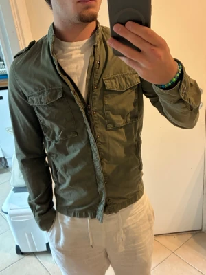 Olivgrön fältjacka  - Säljer en olivgrön overshirt i bomull med två stora bröstfickor och axelklaffar. Overshirten har en avslappnad passform och stängs med knappar framtill. Perfekt att slänga över en t-shirt för en casual look. Bra skick, storlek XS