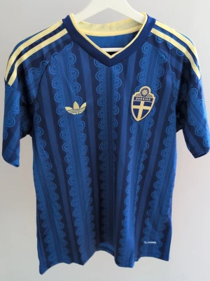 Blå Sverige fotbollströja Adidas -  blå fotbollströja från Adidas med Sveriges landslagsmärke på bröstet och gula detaljer på axlarna. Tröjan har korta ärmar, V-ringning och ett unikt mönster i olika blå nyanser. Materialet är lätt och andas, perfekt för träning eller match.
