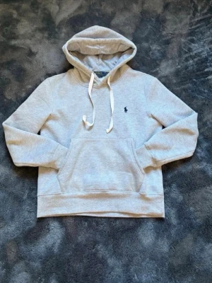 Grå hoodie från Polo Ralph Lauren - Snygg grå hoodie från Polo Ralph Lauren med klassisk huva och vita dragsnören. Tröjan har en stor magficka och den ikoniska mörkblå broderade loggan på bröstet. Tillverkad i mjukt sweatshirtmaterial som är riktigt skönt att ha på sig.