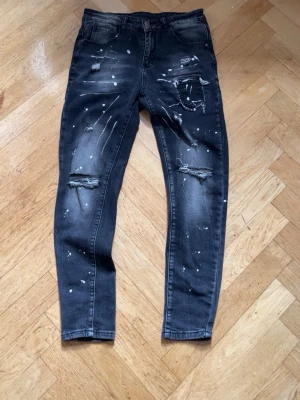 Svarta skinny jeans med slitningar - Säljer ett par svarta skinny jeans med slitningar och vita färgstänk. Jeansen har klassisk femficksmodell, dragkedjegylf och är tillverkade i stretchigt denimtyg för en bekväm passform. Perfekt för en trendig och avslappnad stil.