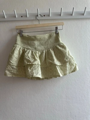 Ljusgul volangkjol med broderi - Supersöt ljusgrön kortkjol med volanger och broderade detaljer. Kjolen har smock i midjan för skön passform och är perfekt för varma dagar. Den luftiga bomullen och de fina broderierna är perfekt för sommaren. 