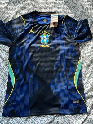 Brasilien matchtröja blå Nike - Snygg blå och svart Brasilien matchtröja från Nike med gula detaljer och CBF-logga på bröstet. Tillverkad i lätt och ventilerande funktionsmaterial, perfekt för fotboll eller träning. Unik design med mönstrade inslag och korta ärmar.