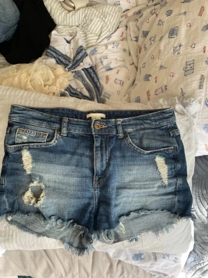 Blå denimshorts med slitningar - Säljer ett par blå jeansshorts med råa kanter och slitna detaljer framtill och baktill. Klassisk femficksmodell med knapp och dragkedja. Perfekta för sommaren och ger en avslappnad vibe.