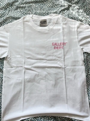 Vit t-shirt från Gallery Dept. - Vit t-shirt från Gallery Dept. med rosa tryck fram och stort tryck på ryggen där det står 'GALLERY DEPT. HOLLYWOOD, CA.'. Klassisk passform och rund halsringning. Perfekt för en clean och trendig look.