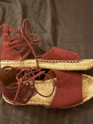 Vinröda espadrillesandaler med snörning - Säljer ett par vinröda sandaler med snörning och dragkedja bak. Sandalerna har flätad sula och öppen tå. Snygg cut-out design på sidorna och trendig espadrillestil. Perfekta för sommaren och ger en bohemisk vibe.