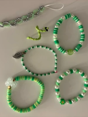 Gröna pärlarmband med berlocker - Säljer ett set med fem handgjorda armband i olika nyanser av grönt och vitt. Armbanden är gjorda av plastpärlor och har detaljer som en silverfärgad bladberlock, en genomskinlig fjäril och små smiley- och blompärlor. Perfekta för att ge din stil en färgglad och lekfull touch.