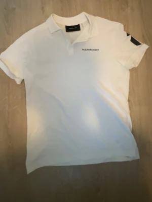 Vit piké från Peak Performance - Klassisk vit pikétröja från Peak Performance med korta ärmar och liten logga på bröstet. Tröjan har krage och knappar vid halsen. Perfekt till en clean och sportig stil.