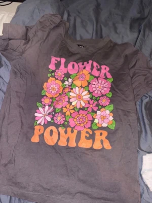 Grå Flower Power t-shirt med blommor - Snygg grå t-shirt med färgglatt tryck av blommor och texten 'Flower Power' i rosa och orange. T-shirten har korta ärmar och en avslappnad passform. Perfekt för dig som gillar retrostil och färgglada motiv.