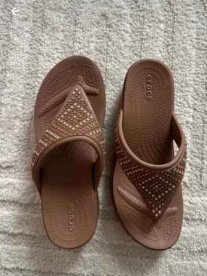 Bruna Crocs sandaler med strass - Snygga bruna sandaler från Crocs med breda remmar dekorerade med glittrande strass i geometriskt mönster. Tillverkade i mjukt och bekvämt syntetmaterial med ergonomisk sula för extra komfort. Perfekta för dig som vill ha både stil och bekvämlighet.