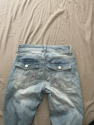Ljusblå jeans med ficklock från Nelly - Säljer ett par ljusblå jeans med hål på knäna och dubbla knappar i midjan. Jeansen har bakfickor med ficklock och knappdetaljer. Utsvängda och lågmidjade. Perfekta för en avslappnad och trendig look. Säljer då jag ej använder dom. De enda defekterna de har är att det finns typ sminkfläckar eller liknande framtill runt knäområdet. Syns på bilderna. De kanske går bort med något bra tvättmedel men inget jag verkligen försökt o få bort. Annars är de i bra skick.