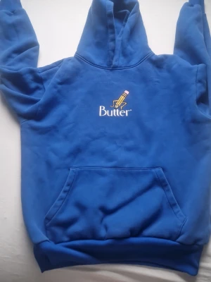 Blå hoodie från Butter med tryck - Blå hoodie från Butter med huva och stor känguruficka. På bröstet finns ett tryck med en smörpinne och texten 'Butter' i vitt. Hoodien är gjord i mjukt fleece-material och har ribbade muddar vid ärmar och nederkant. Perfekt för dig som gillar streetwear och vill sticka ut.