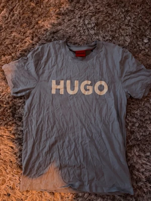 Grå HUGO t-shirt med tryck - Snygg grå t-shirt från HUGO med stort vitt logotryck på bröstet. Klassisk rund halsringning och korta ärmar. Tillverkad i mjuk bomull för en skön känsla. Perfekt till jeans eller shorts för en clean och enkel stil.